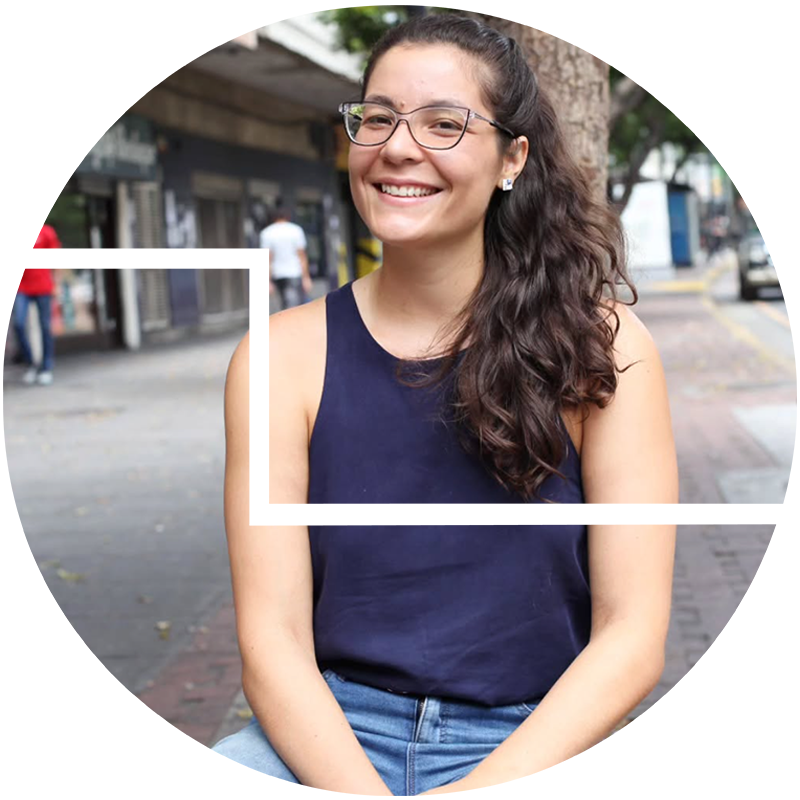 Keyla Brando with dark, curly hair and glasses, smiling and looking to the side, seated outdoors in an urban setting.